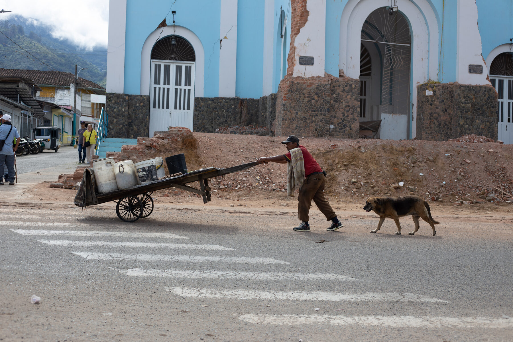 A man pulling a hand cart loaded with water containers past the damaged church, a dog walking alongside